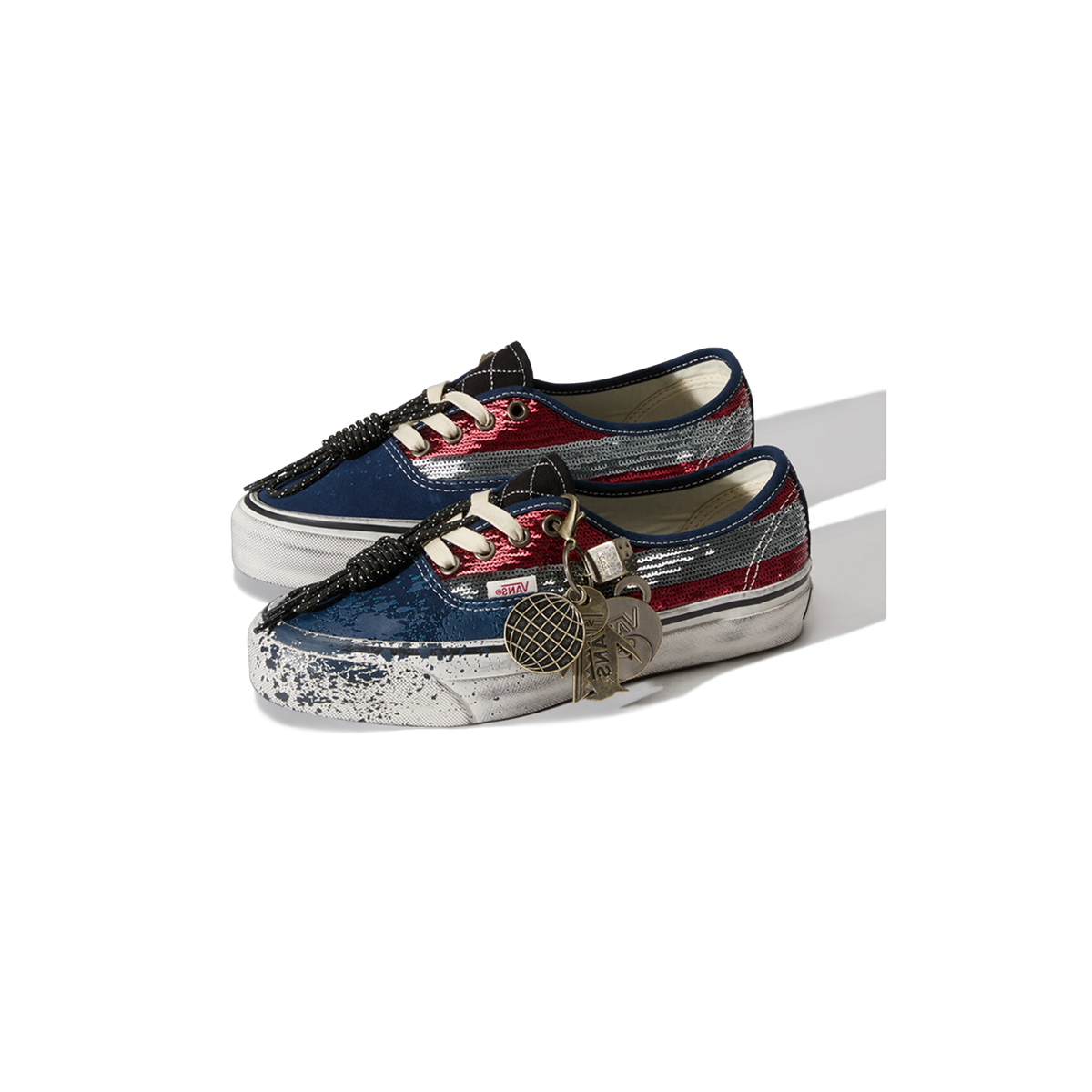 Pair of Vans Authentic sneakers with red and silver sequined stripes, navy toe and heel, distressed paint-splattered white sole, and metal charms hanging from the black paracord laces.