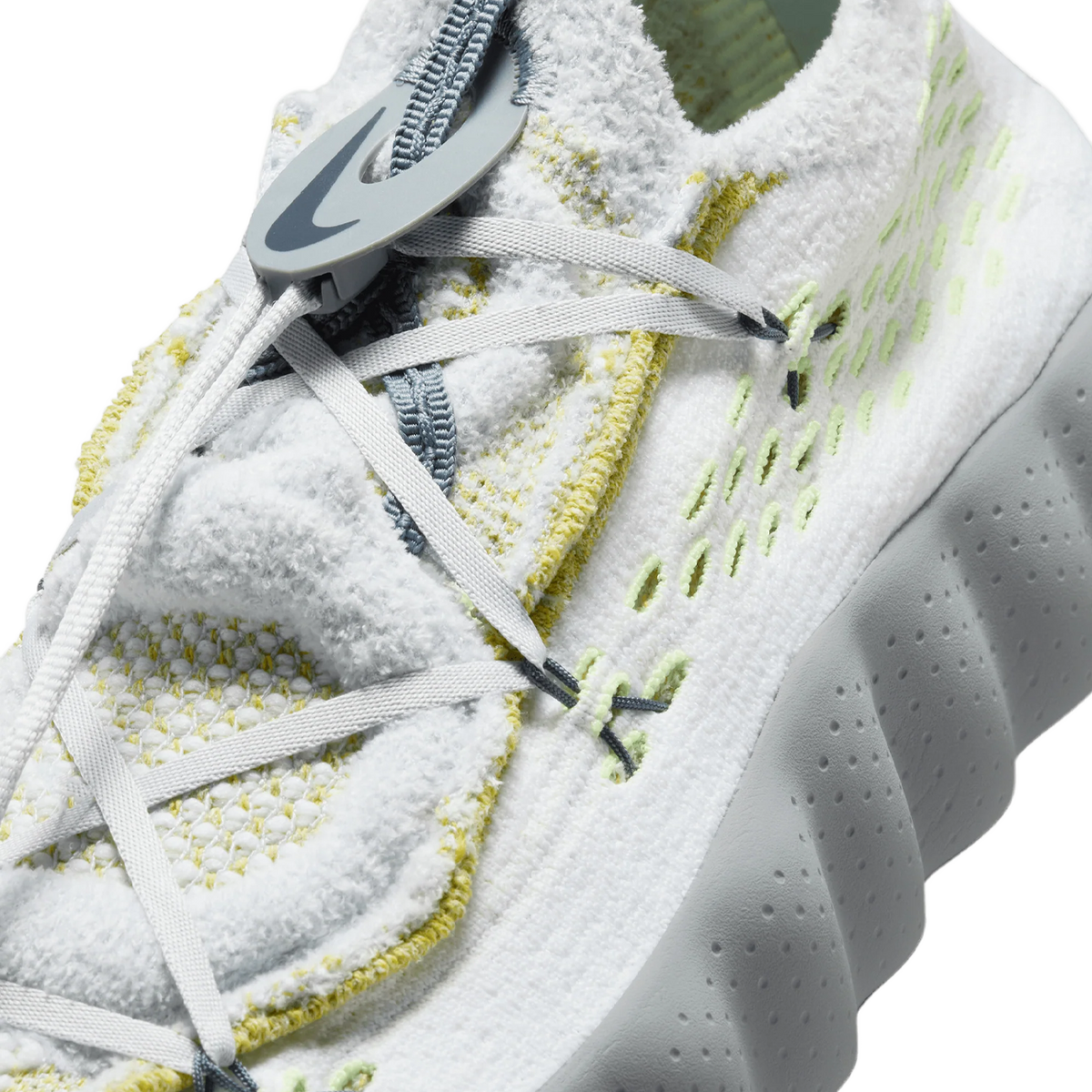 Close-up of a white Flyknit sneaker's lacing area showing a grey Nike swoosh pull tab, grey laces, and volt-colored stitching accents.