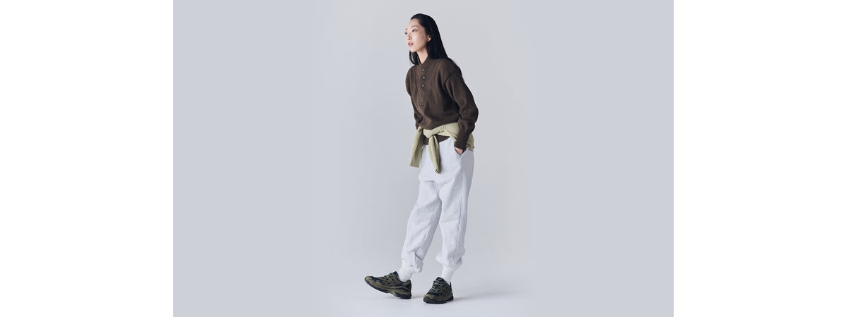 A woman wearing grey sweatpants and a brown sweater stands modeling green and grey Asics Gel-NYC sneakers.