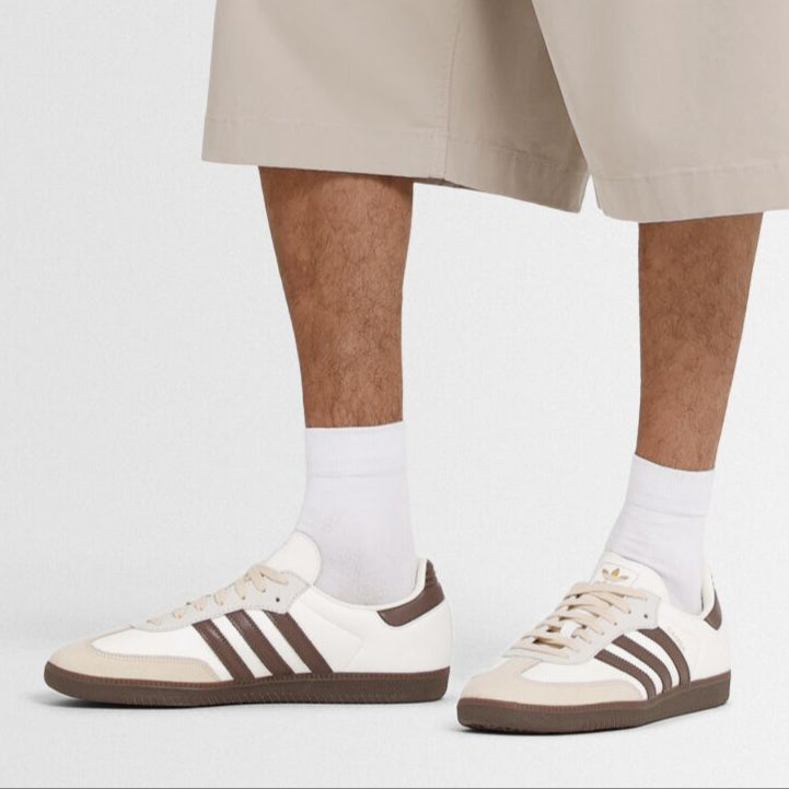 A person wearing white socks and shorts models a pair of white Adidas Samba sneakers with brown stripes and gum soles.
