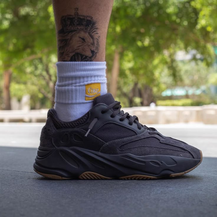 Person wearing a black Adidas Yeezy Boost 700 Utility Black sneaker on foot with a white sock, shown outdoors with trees in the background