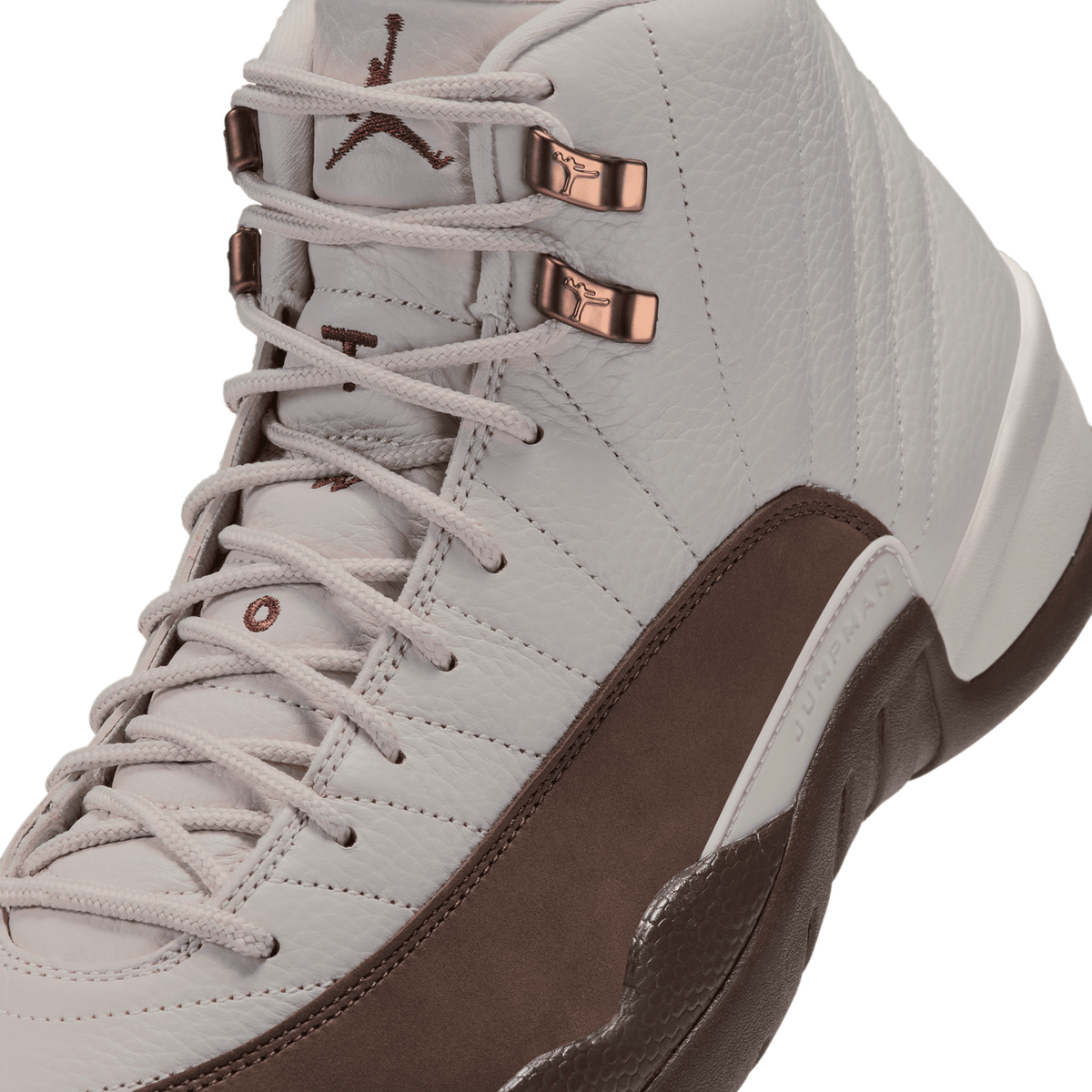 Close-up angled view of a white Air Jordan 12 with brown toe box and heel, metal eyelet lace hooks, and embroidered Jumpman logo on the tongue