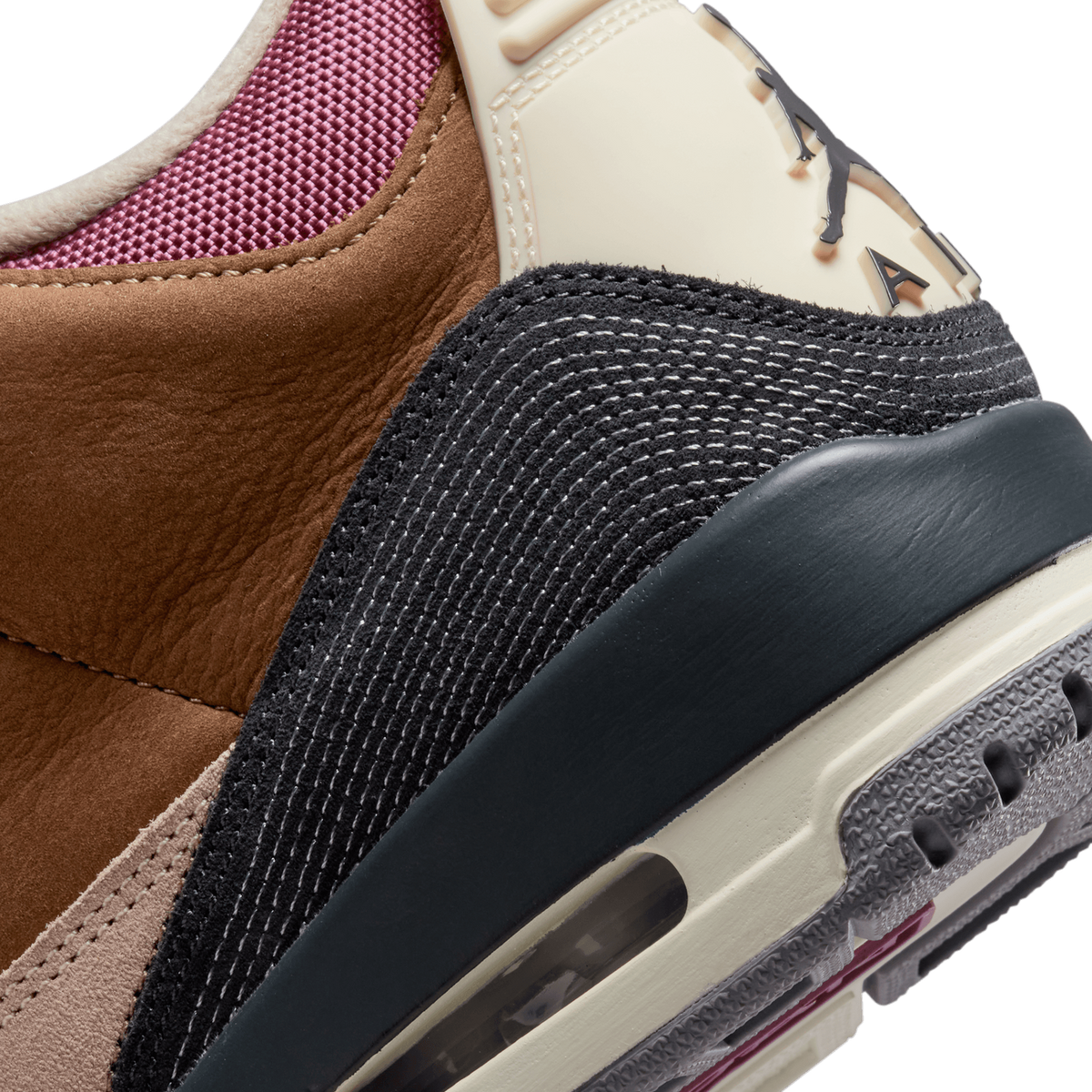 Extreme close-up of the heel area of a brown, beige, and black high-top sneaker, showing brown nubuck, black textured panels with contrast stitching, a beige heel tab with a dark Jordan Wings logo, a dark rubber heel collar, and a pink mesh lining vi