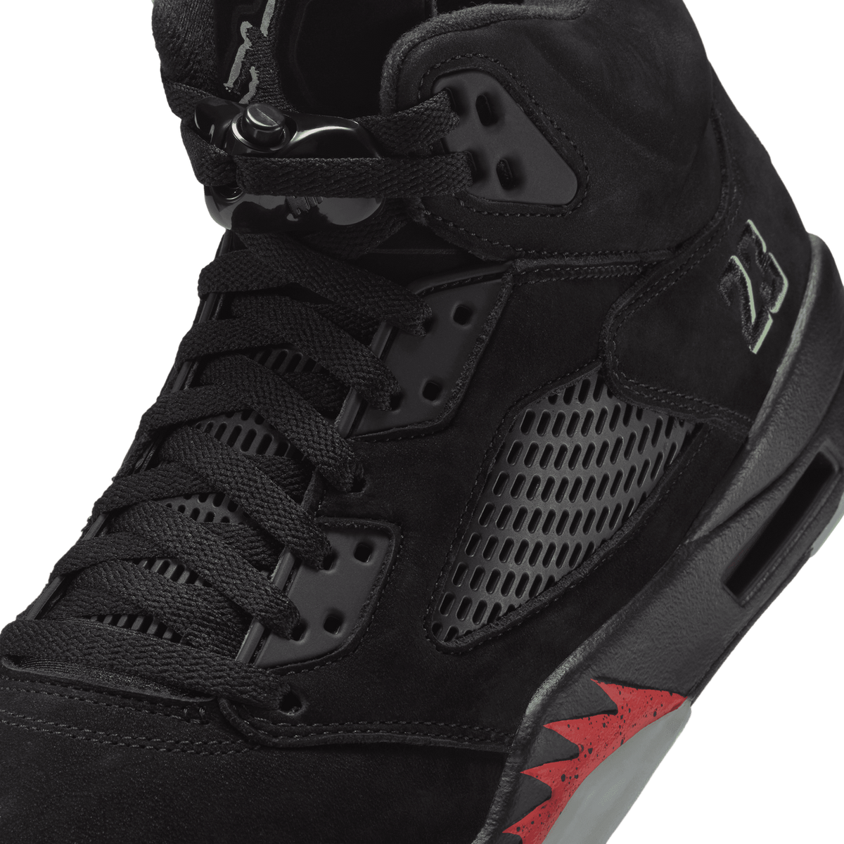 Close-up view of a black suede Air Jordan 5 sneaker showing the lace eyelets, metal lace lock, mesh tongue panel, and red translucent midsole detailing.