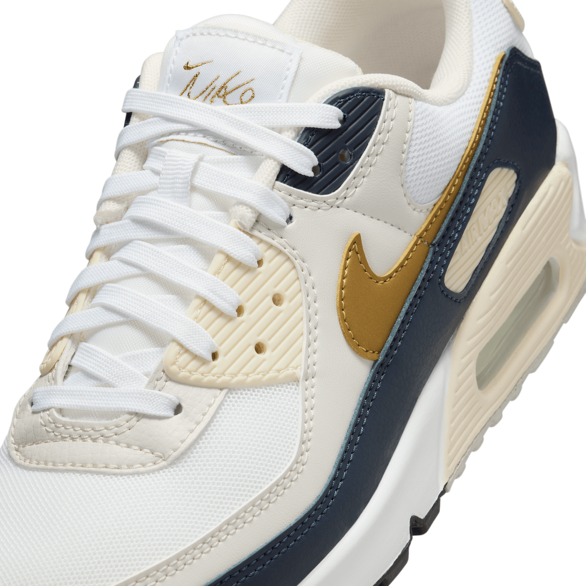 Close-up of a white Nike Air Max 90 sneaker showing the gold Swoosh logo, navy heel panel, and signature-style Nike script on the tongue tag.