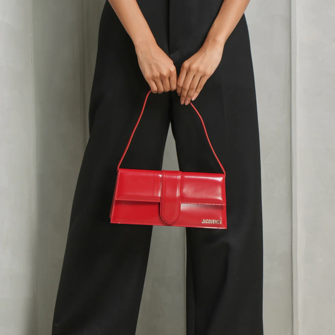 Red jacquemus handbag held by a person wearing black pants against a neutral background