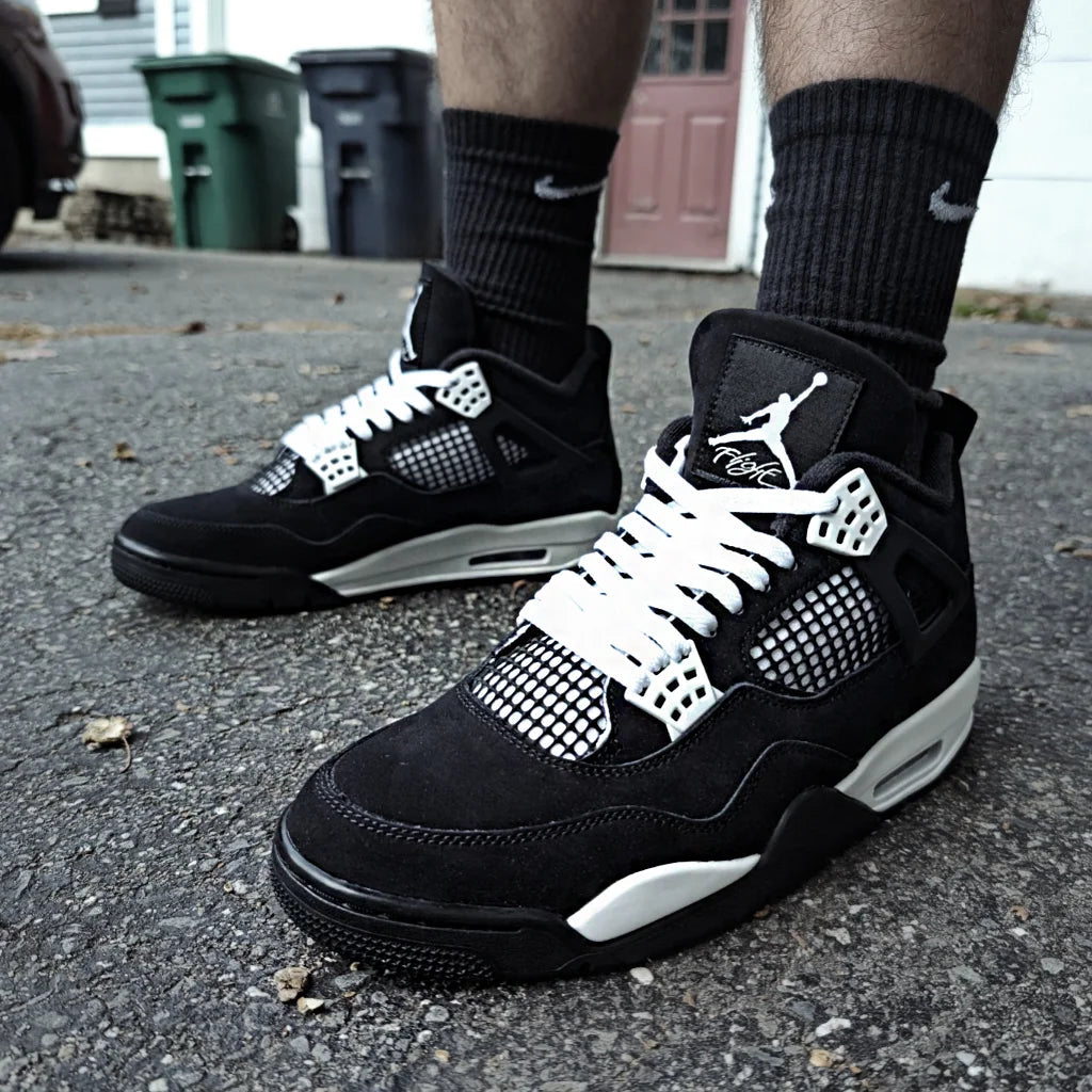 A person wears black suede Jordan 4 Retro sneakers with white mesh side panels, white laces, and white midsoles while standing on a paved driveway.
