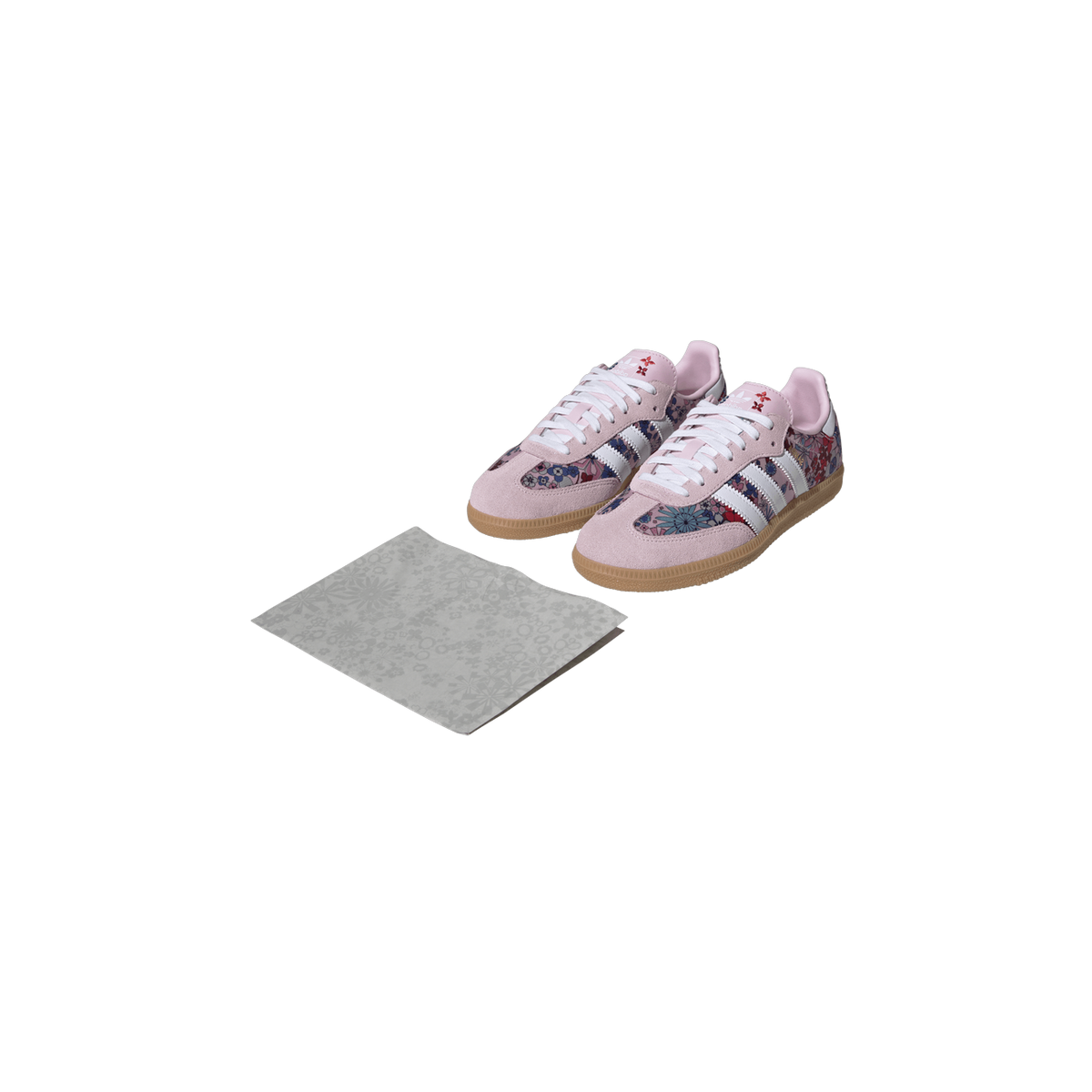 Pair of pink Adidas Samba sneakers with floral multicolor print, white stripes, and gum soles, shown beside a matching floral cloth.