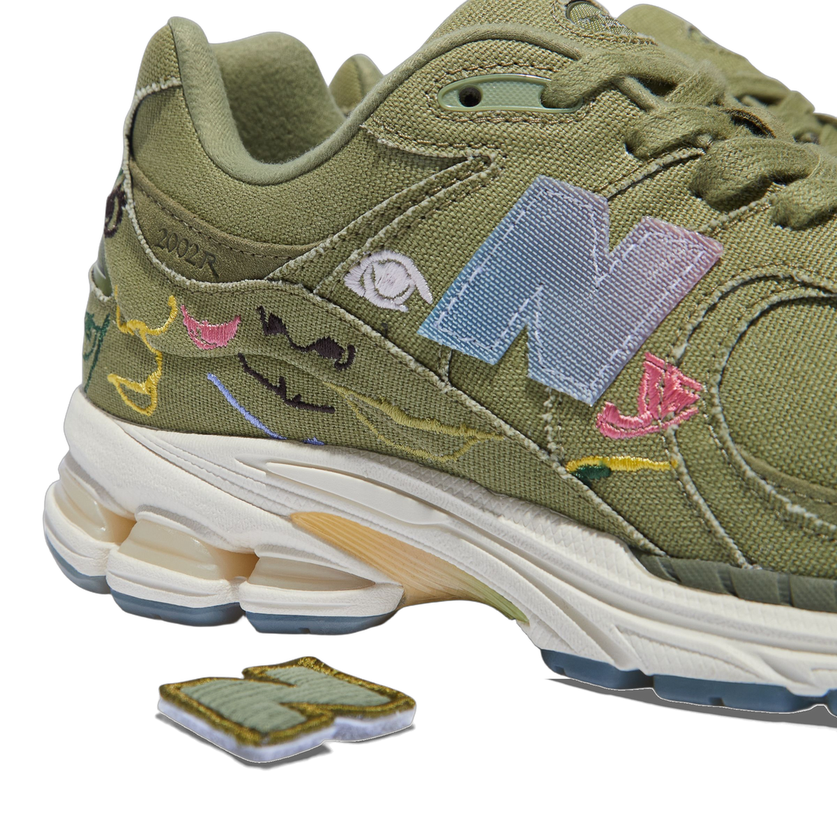 Close-up rear angle of an olive green New Balance 2002R with colorful embroidered face doodles, a detachable interchangeable N logo patch, and a cream and tan midsole.