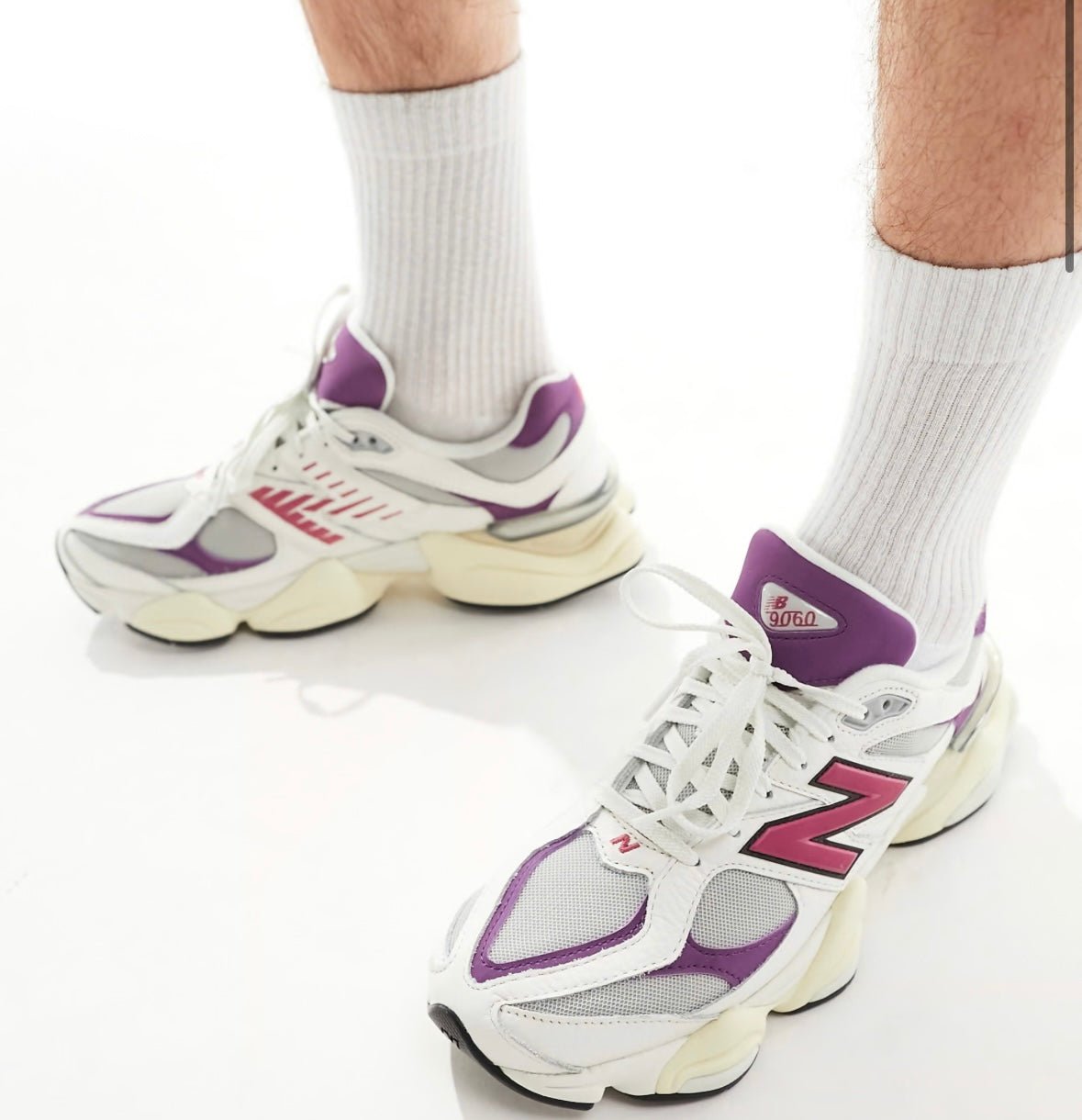 Person wearing white New Balance 9060 sneakers with purple accents and a pink N logo, shown on feet with white socks.