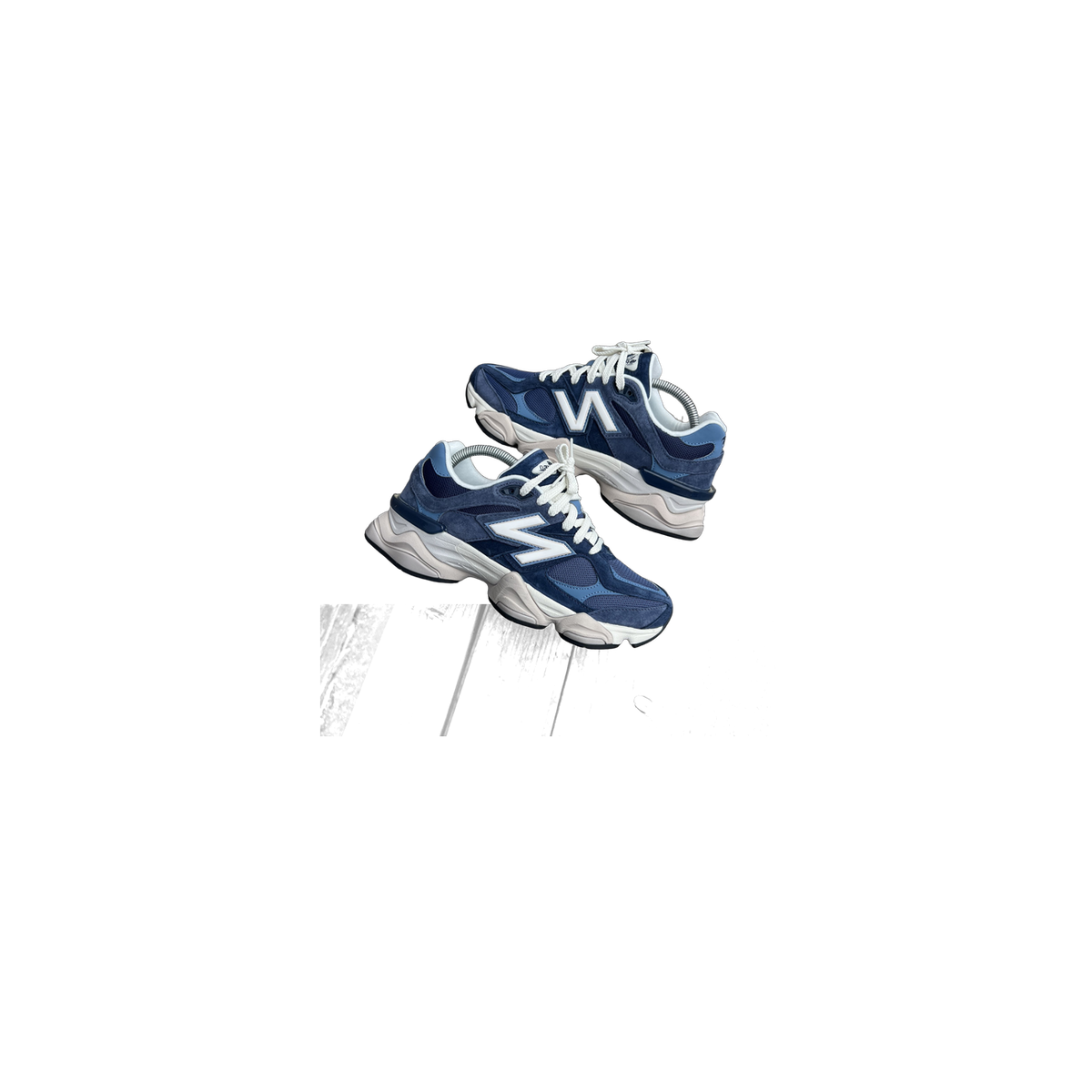 A pair of navy blue New Balance 9060 sneakers with white N logos and chunky off-white soles, resting on a wooden plank.