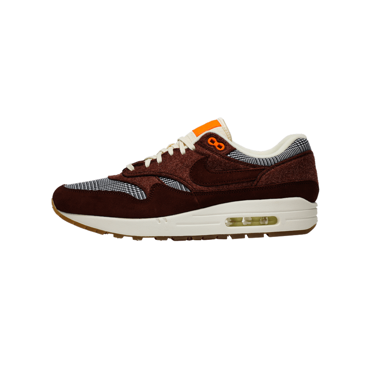 Left Nike Air Max 1 sneaker in dark brown suede with houndstooth panels, orange tongue tab, cream laces and midsole, shown from the side with toe pointing left.