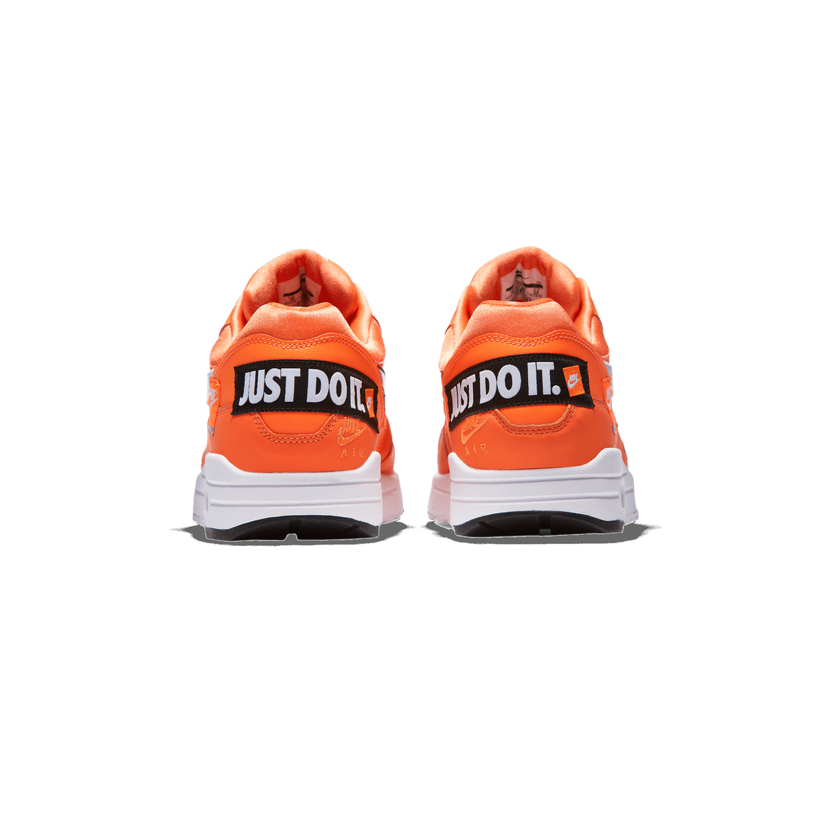 Rear view of a pair of orange Nike Air Max 1 sneakers on a white background, showing the heel tabs with black 'JUST DO IT.' woven labels, white midsoles and black outsole edges.