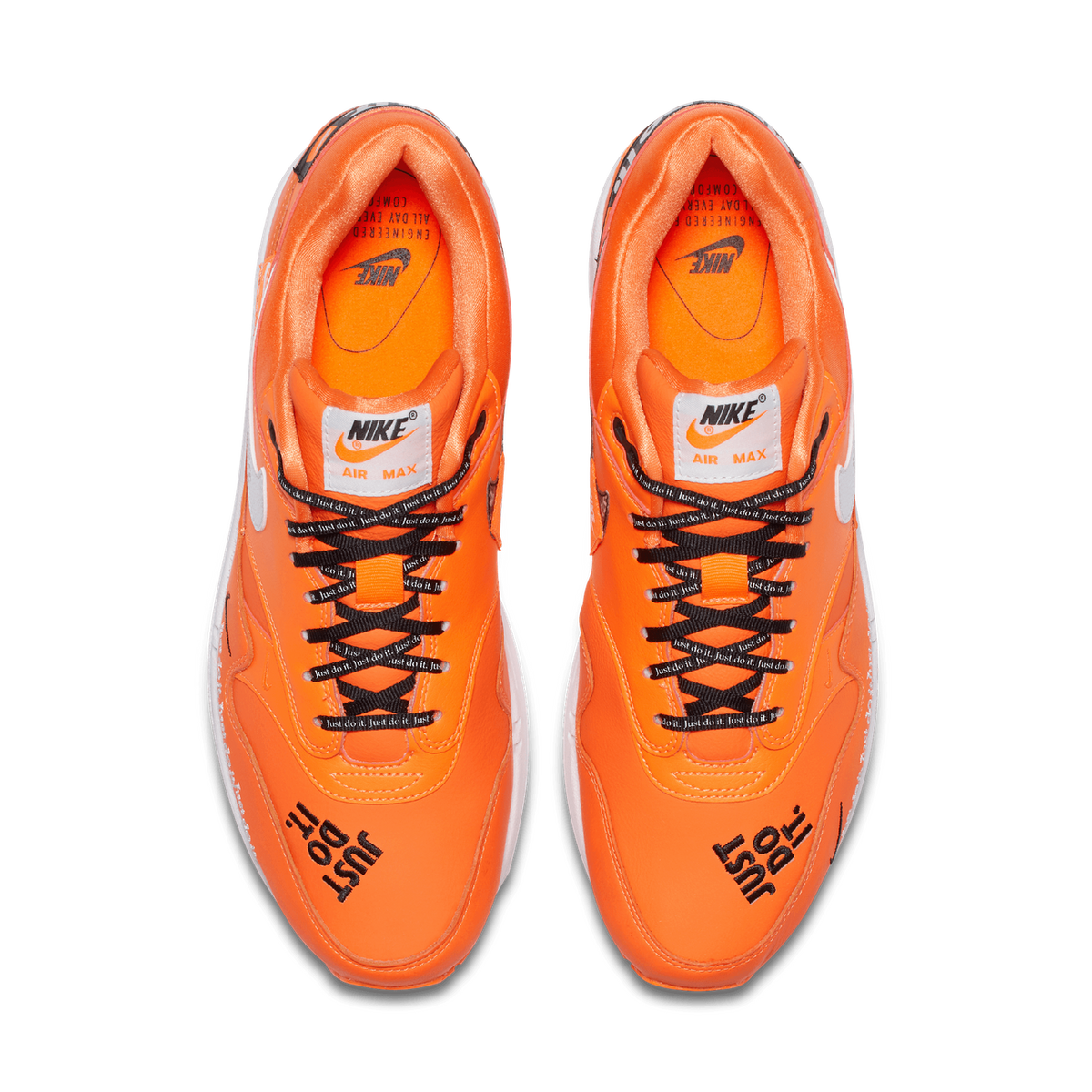 Top-down view of a pair of orange Nike Air Max 1 sneakers placed side by side with toes pointing toward the bottom of the frame, showing black laces with 'Just do it' text, white Swoosh logos, black embroidered 'JUST DO IT' lettering on the toe boxes