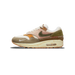 Outside view of a left Nike Air Max 1 sneaker with cream, olive green, pink, orange and brown panels, an olive Swoosh, red cross-stitch detailing, and a crackled cream midsole with visible Air unit.