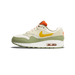 Side profile of a Nike Air Max 1 sneaker with the toe pointing left, showing cream and white upper panels, a yellow Swoosh, olive-green midsole base and toe cap, orange heel tab, and a visible orange Air unit in the heel.