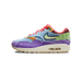 Left Nike Air Max 1 sneaker in side view with denim blue upper, purple and green paisley panels, red laces, and a visible Air unit in the cream midsole