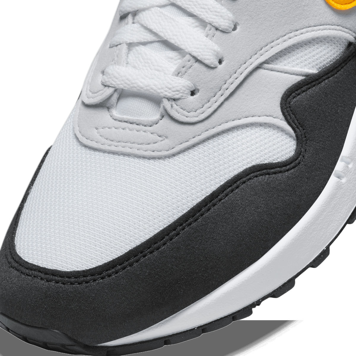 Close-up detail of the toe box and lacing of a white and black Nike Air Max 1 with a gold swoosh logo visible at the top edge, showing white mesh, grey overlays, black suede panels, and a white midsole.