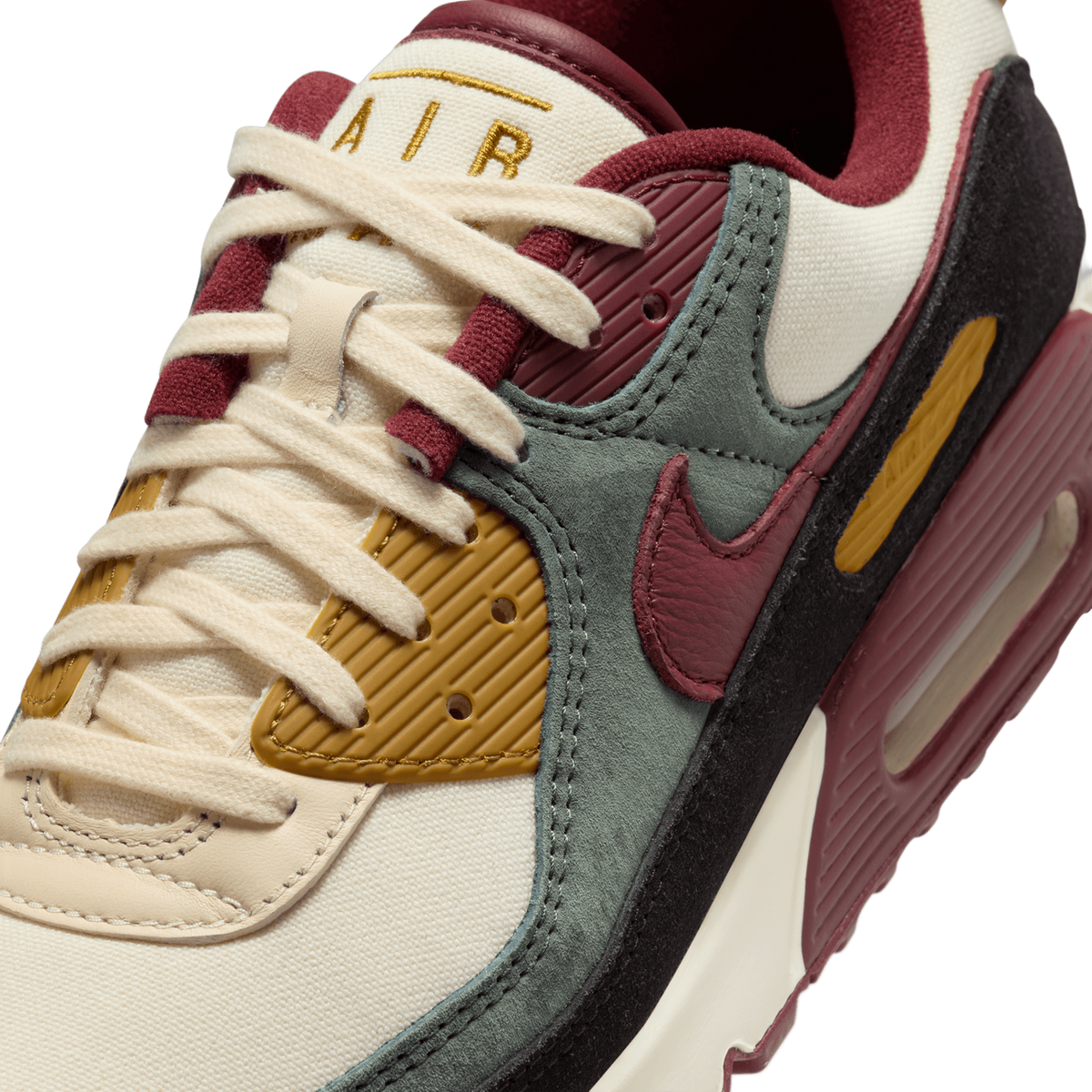 Close-up angled view of a Nike Air Max 90 showing the cream laces, embroidered 'AIR' text, and swoosh in burgundy, sage green, mustard, and black panels.