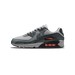 Left Nike Air Max 90 sneaker in grey and black with crackle texture, orange Air Max branding, and visible red-tinted air unit, shown from the side with toe pointing left.