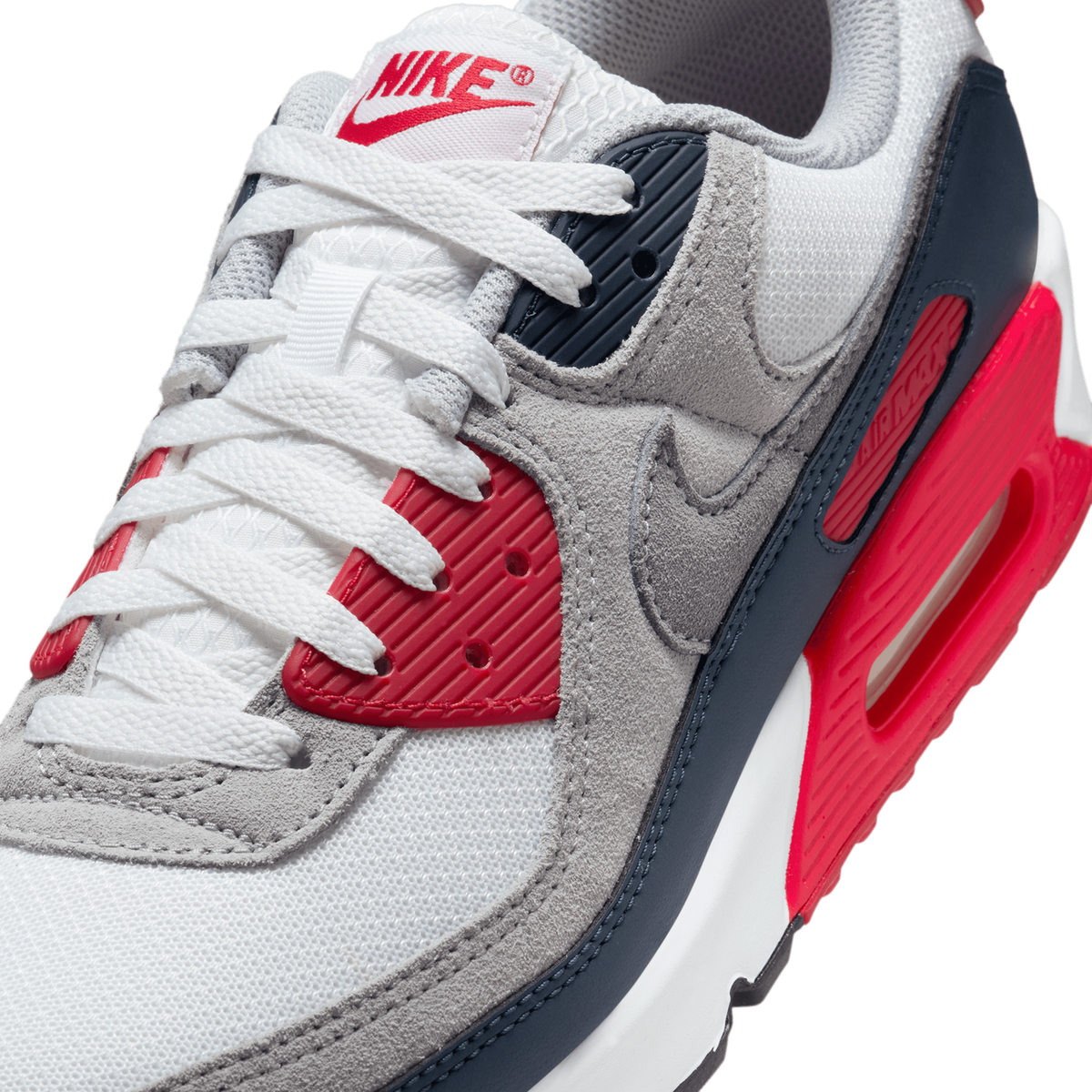 Close-up angled view of a white, grey, navy and red Nike Air Max 90 showing the laces, tongue with red Nike logo, swoosh, and visible Air unit in the midsole.