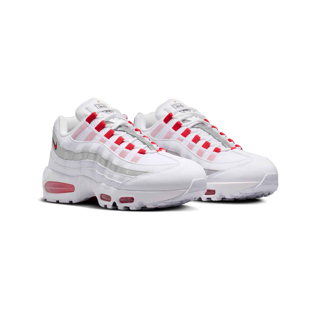 Front-angled view of a pair of white Nike Air Max 95 SE sneakers with red pull tabs, silver layered overlays, red visible air units, and a small crest emblem on the tongue.