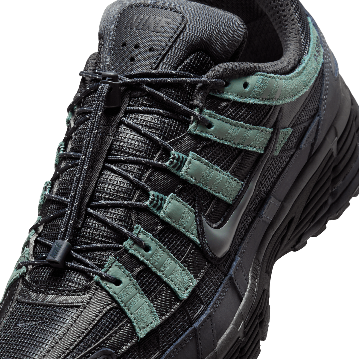 Close-up of a black and grey Nike P-6000 sneaker showing the tongue with a Nike logo, black toggle laces, and sage-green suede overlays with stripe details on the mesh upper.