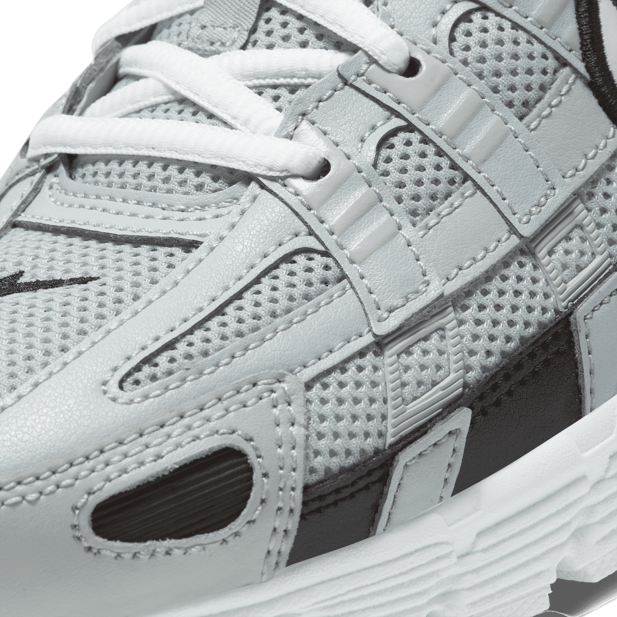 Close-up detail of a silver and white Nike P-6000 sneaker showing mesh and leather panels, white laces threaded through eyelets, black Swoosh embroidery, and the white midsole with ridged texture.