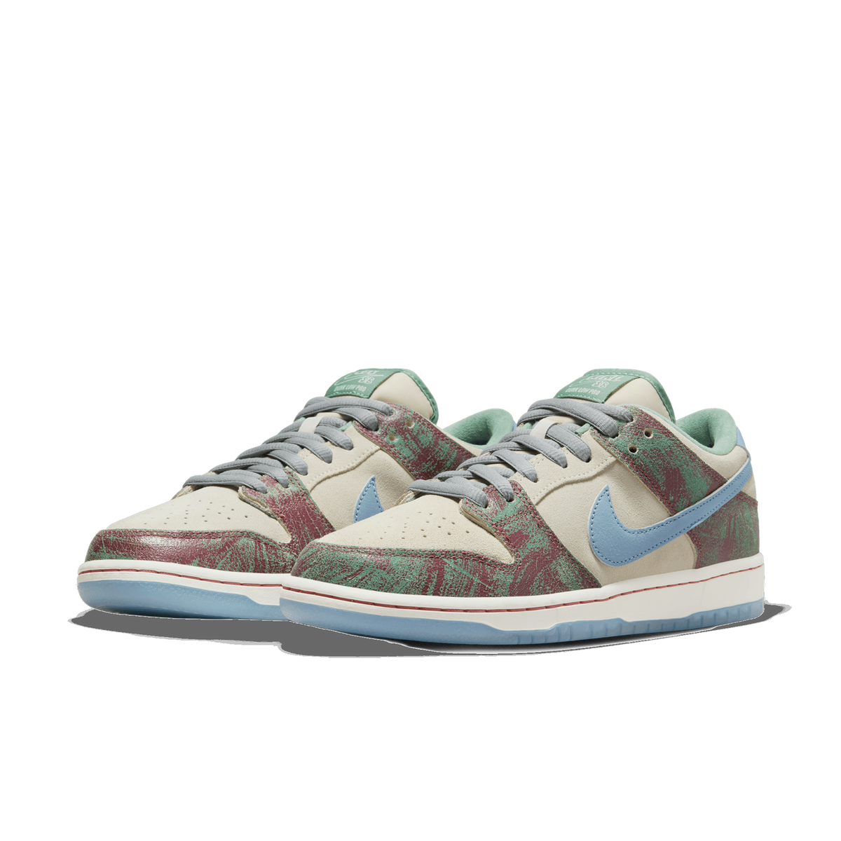 A pair of Nike SB Dunk Low sneakers angled side by side, showing cream suede panels, a mottled green and maroon textured overlay, light blue Swoosh logos, and a cream and light blue sole, with the left shoe's toe pointing left.