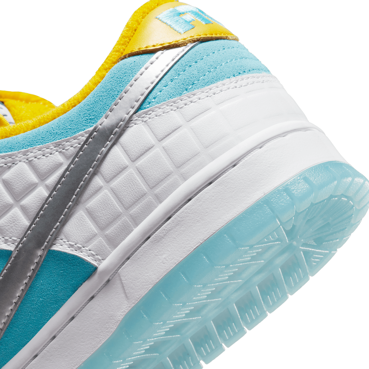 Close-up of the heel and lateral side of a Nike SB Dunk Low shoe showing turquoise suede panels, a white textured midsole with a diamond pattern, a silver swoosh, a yellow tongue pull tab with embossed blue lettering, and a translucent light-blue rub