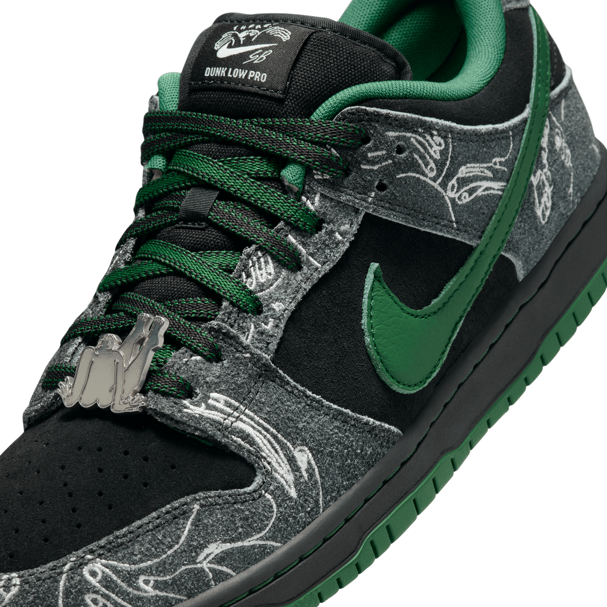 Close-up angled view of a black and grey Nike SB Dunk Low with green Swoosh, laces, and lining, featuring a grey paisley-pattern overlay and a silver figure-shaped lace charm near the toe.