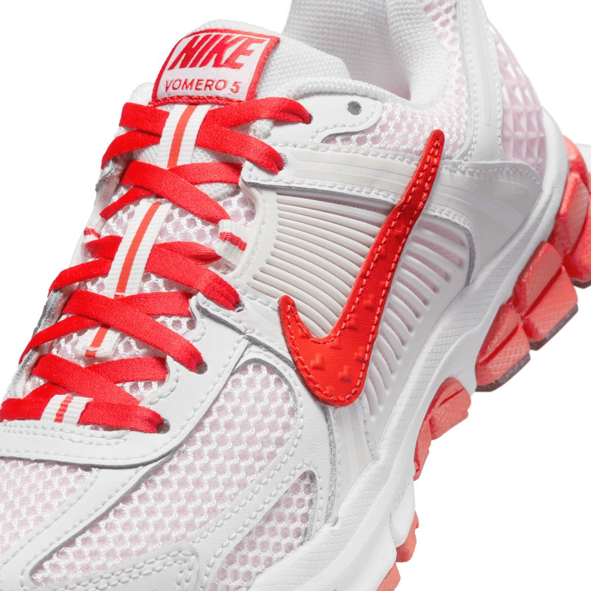Close-up detail of a white and red Nike Zoom Vomero 5 sneaker showing the red woven Nike tongue label reading 'VOMERO 5', red laces, a red Swoosh logo on the side, and coral-red cushioning pods along the heel.