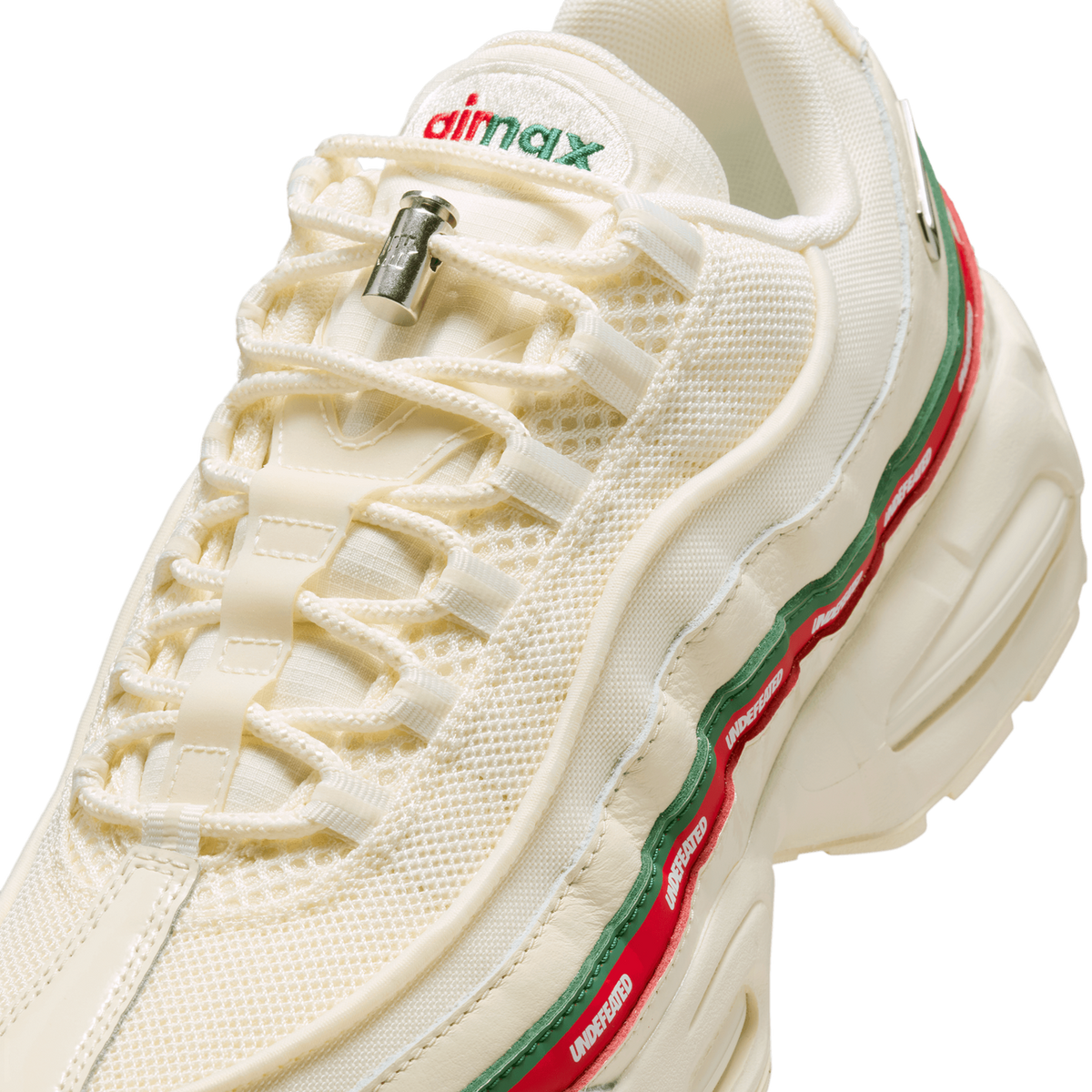 Close-up angled detail of a cream-colored Nike Air Max 95 showing the lacing system with a silver cord lock, an embroidered red and green 'air max' heel tab, and a side stripe with green and red bands printed with white 'UNDEFEATED' text.