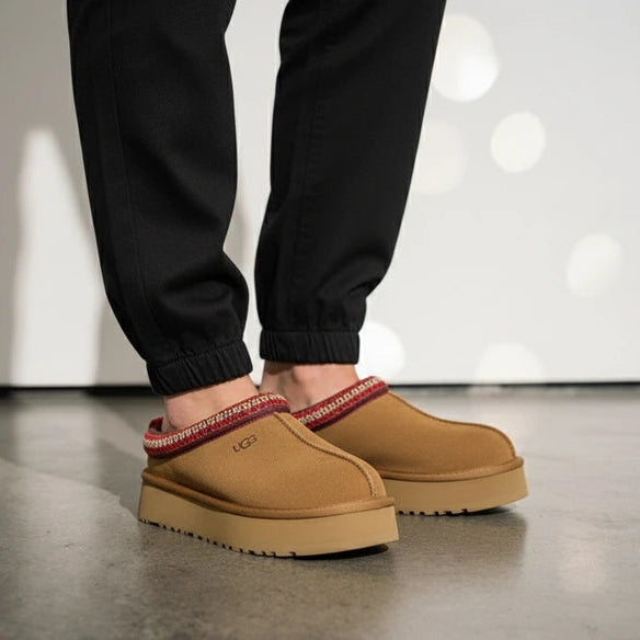 A person wearing black joggers stands in a pair of chestnut brown UGG Tazz slip-on slippers with red and cream patterned trim and a thick tan platform sole.