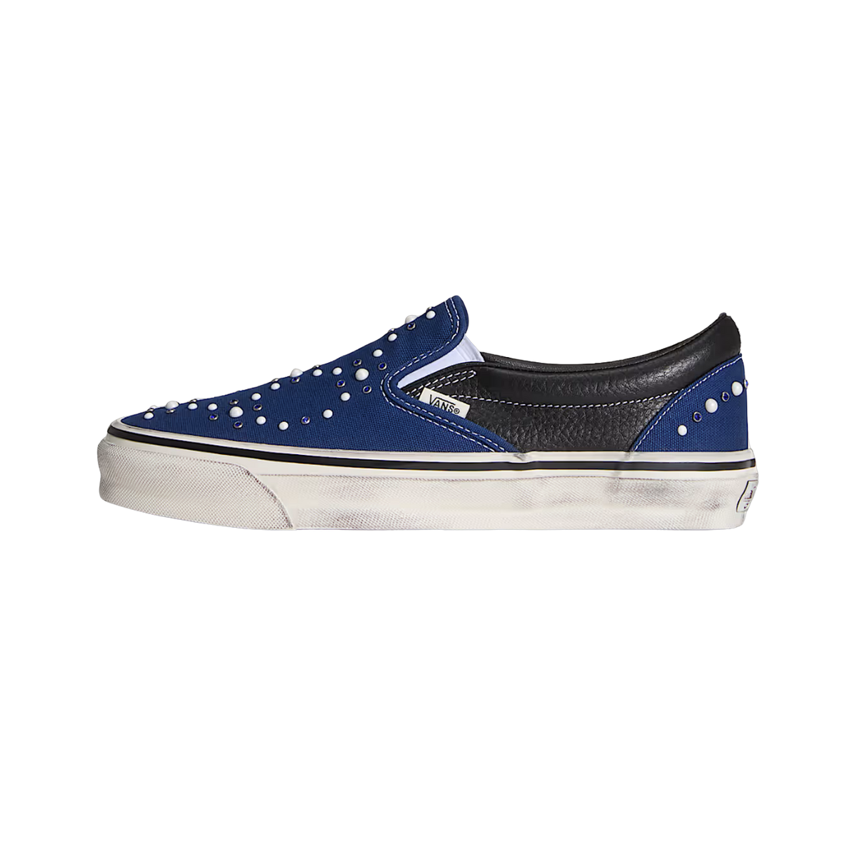 Side view of a blue slip-on sneaker with black leather heel panel, pearl-like bead embellishments across the toe and heel, and a distressed off-white sole.