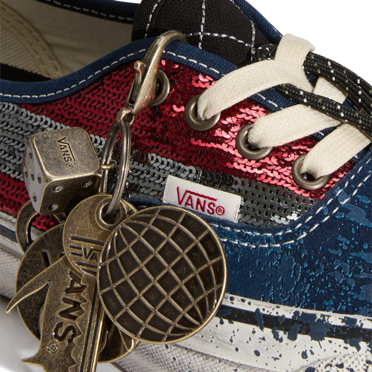 Close-up of a Vans Authentic sneaker's eyelets and tongue with red and grey sequin panels, navy canvas, white laces, a Vans fabric tag, and hanging metal charms including a dice, key, and globe pendant.